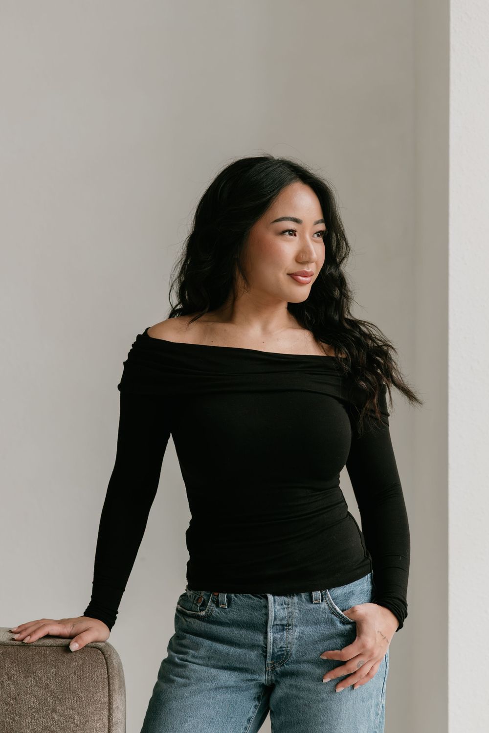 A person in a black off-shoulder top and blue jeans poses against a neutral background in a fashion photography series.