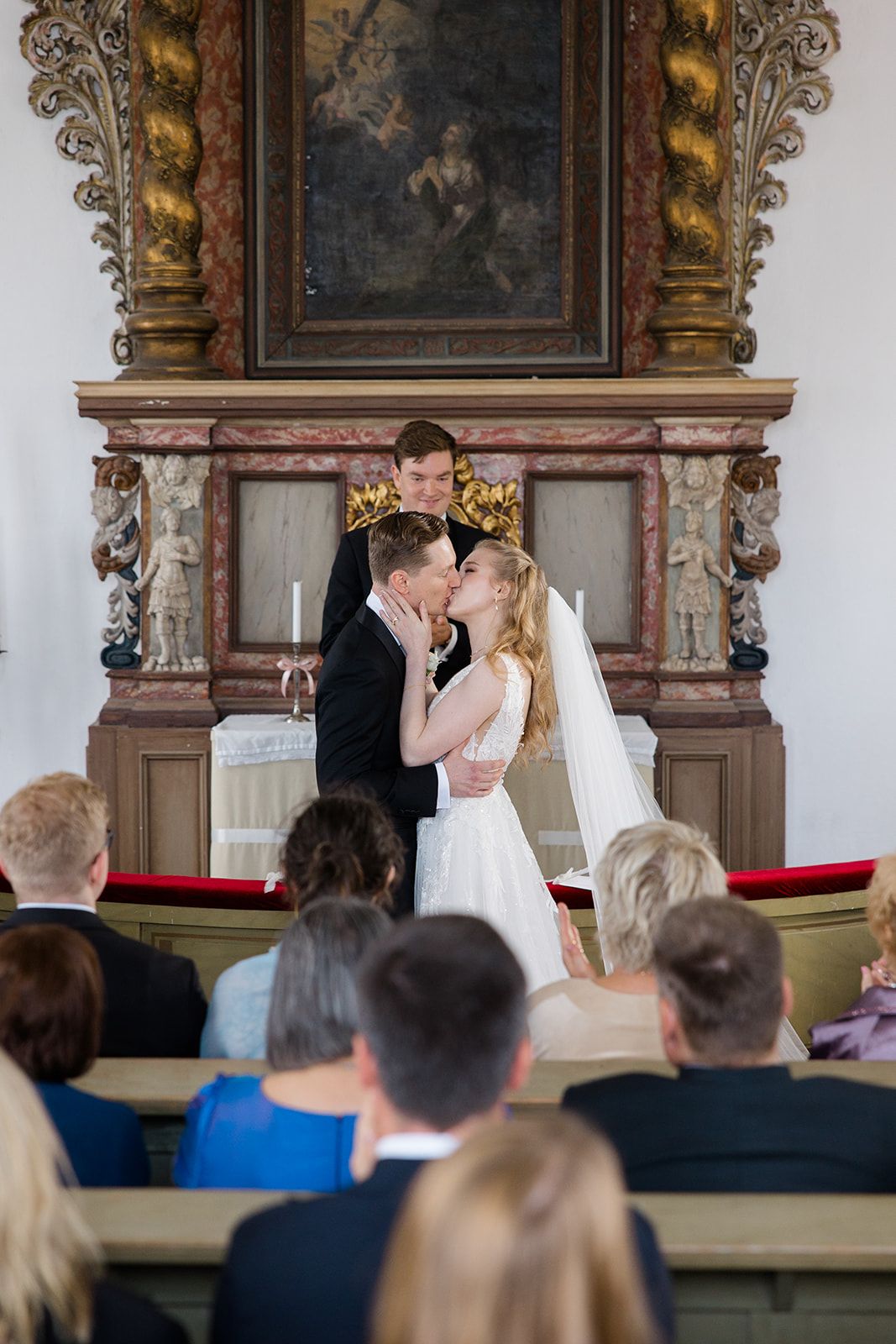 A couple shares their first kiss during a wedding ceremony in an ornate church with guests watching from the pews.
