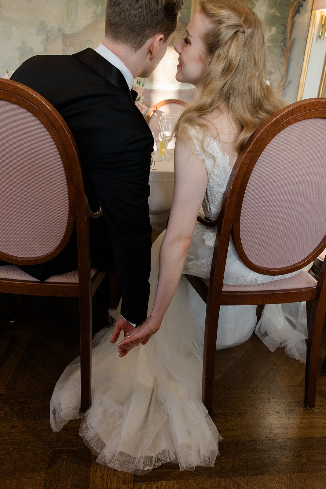A romantic wedding couple shares intimate kisses while seated in antique chairs on a hardwood floor.