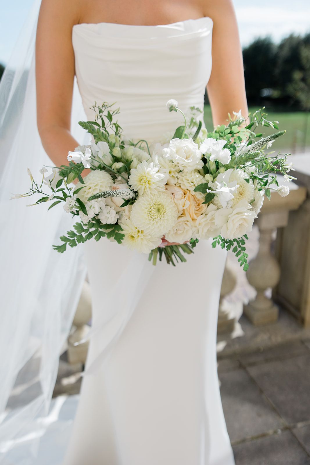 A delicate white bridal bouquet with fresh peonies, dahlias and greenery held against an elegant strapless wedding gown.