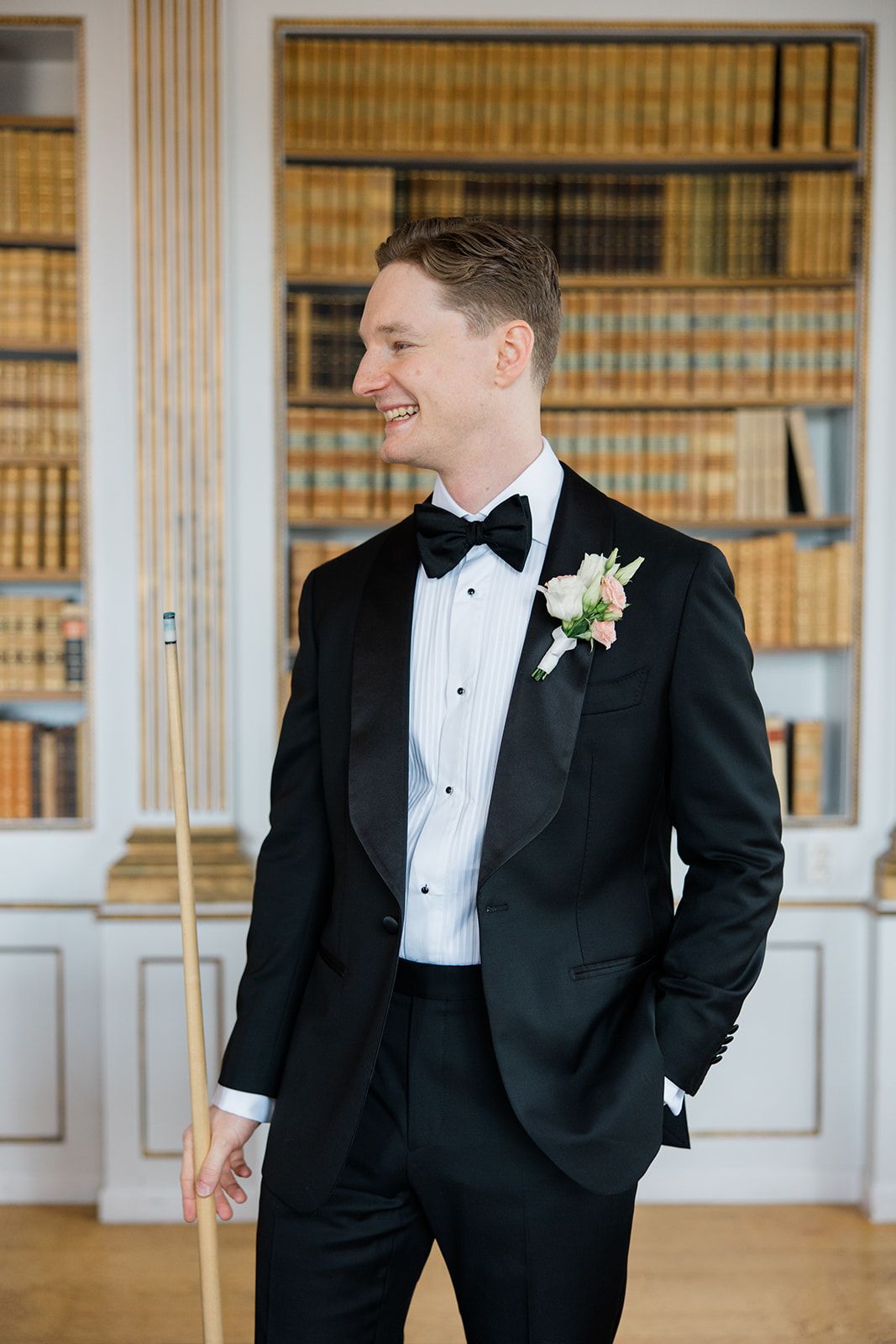 Groom in classic black tuxedo and bow tie stands in elegant library setting with wooden bookshelves.