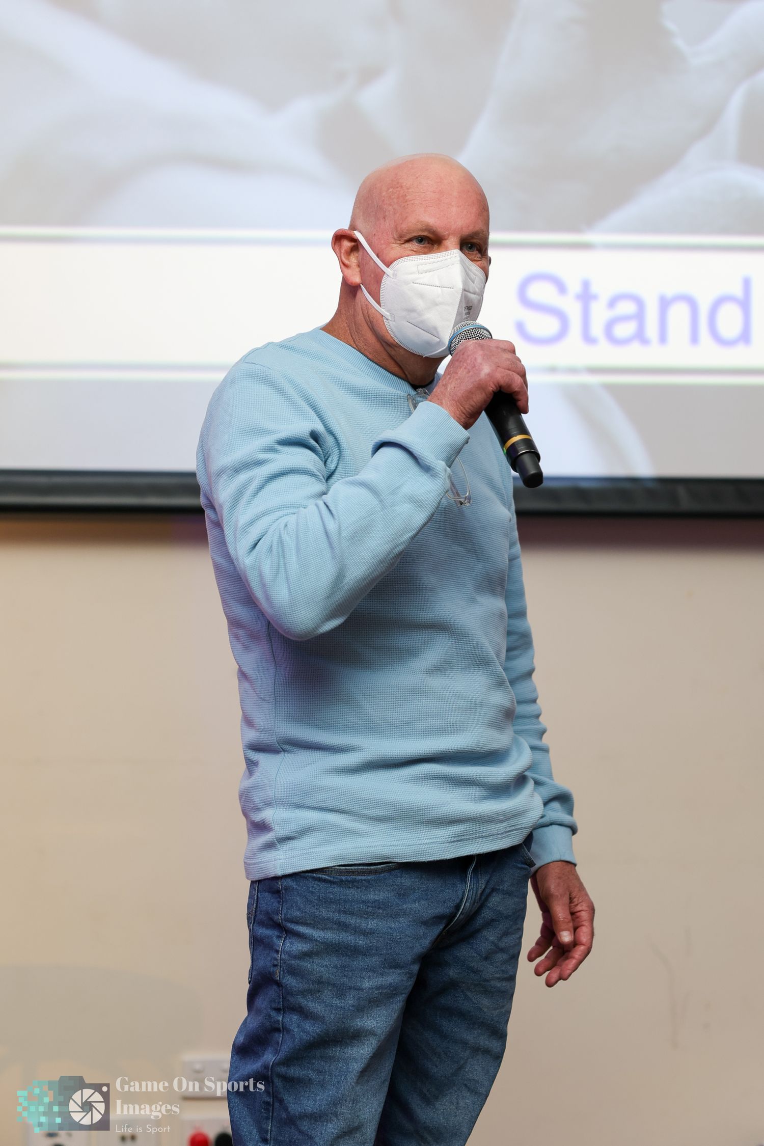A person wearing a light blue sweater and jeans gestures while speaking into a microphone during a presentation. Andrew Smythe.
