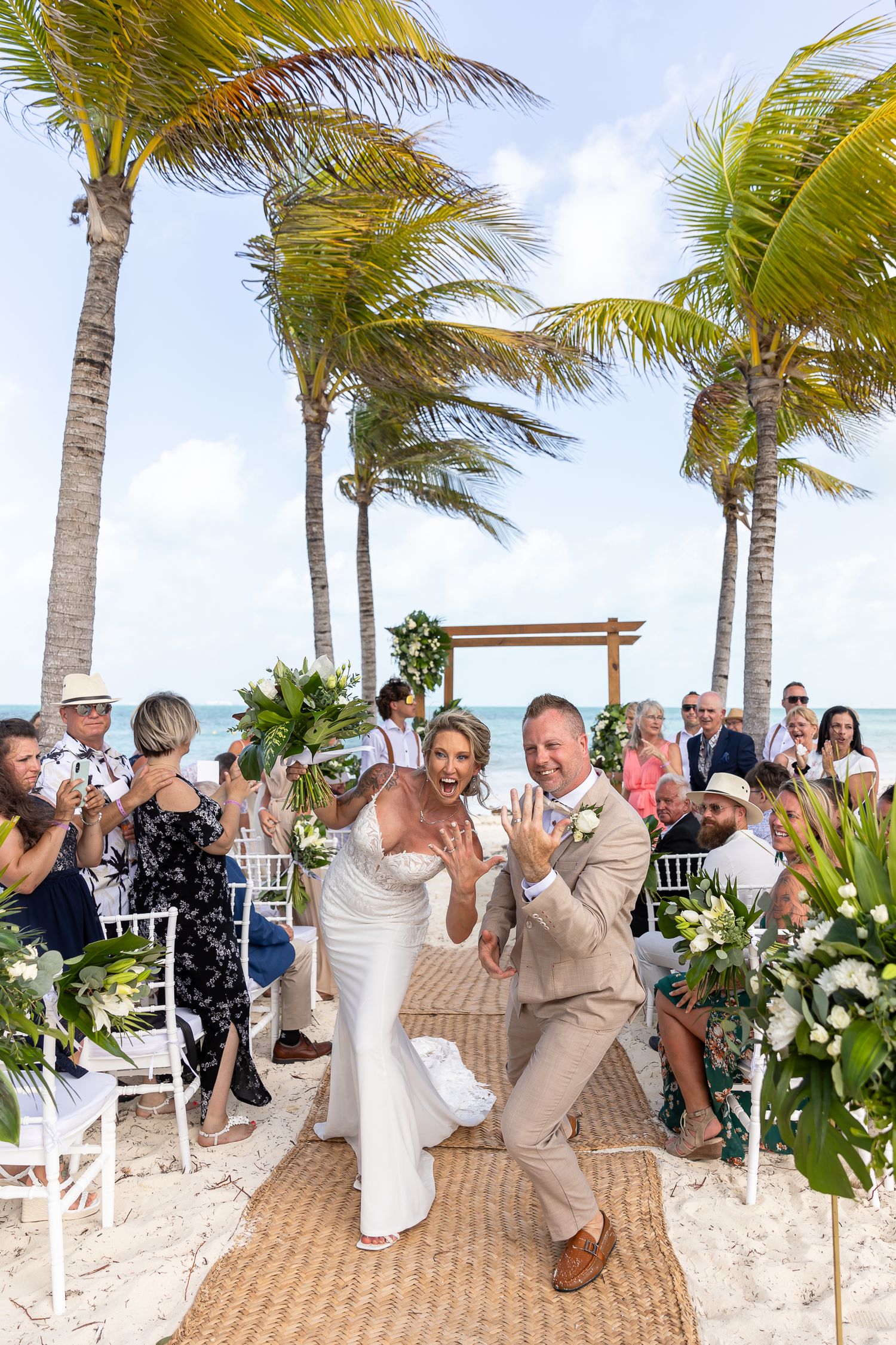Wedding guests on a tropical beach with palm trees laugh and react during a fun candid moment at a destination ceremony.