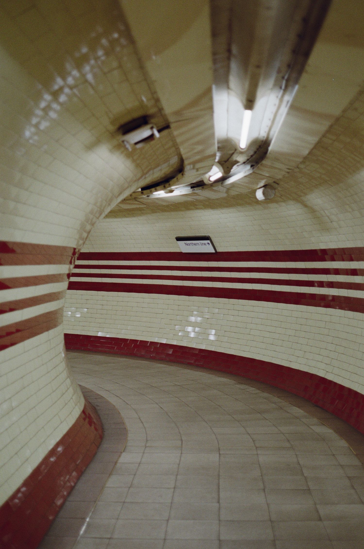A curved subway tunnel corridor with white tiled walls and a red stripe pattern leading around the bend.