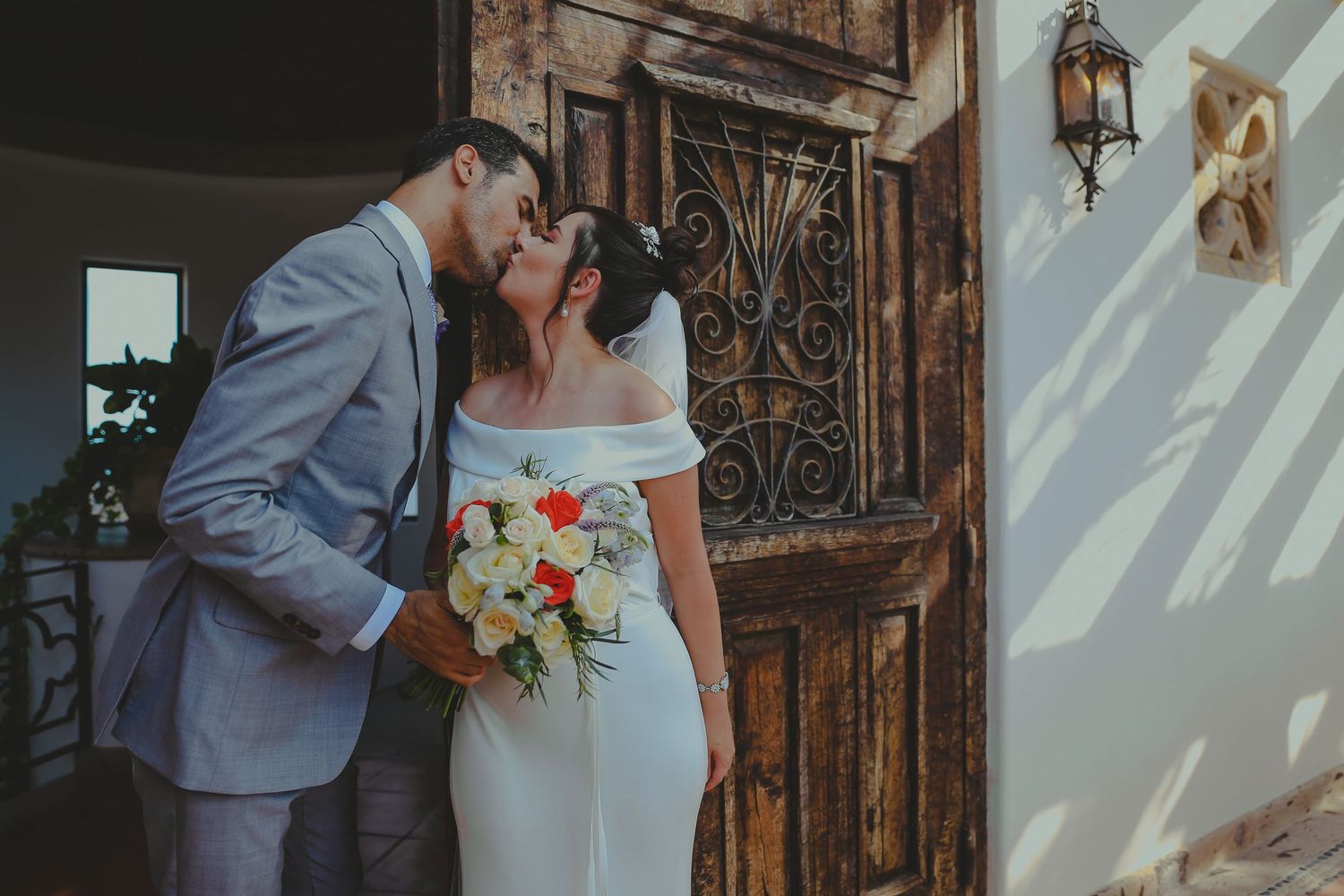 A romantic wedding couple embraces in front of an ornate wooden door at a rustic venue.Pedregal Los Cabos Wedding Photographer