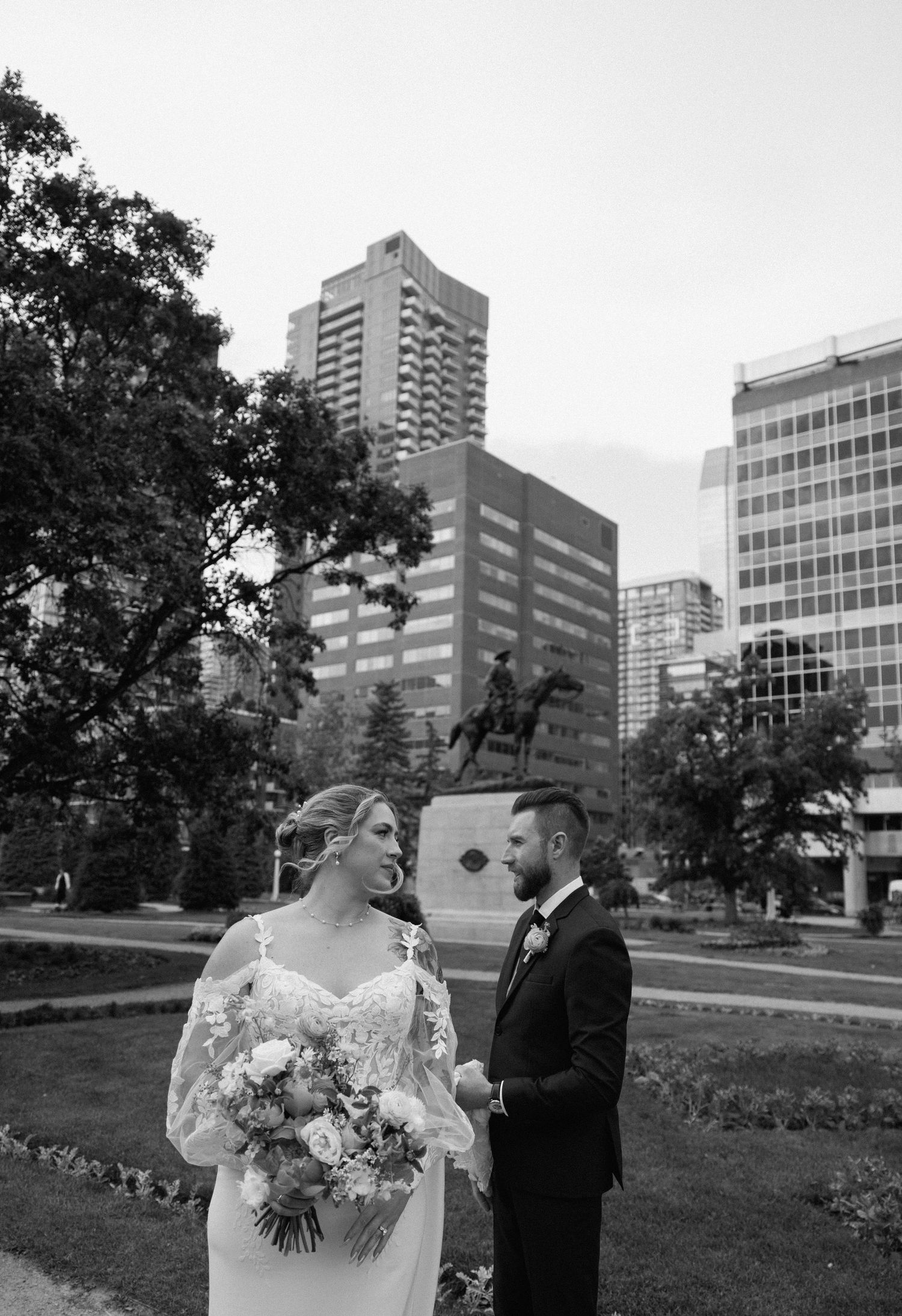 A wedding couple poses outdoors with modern city buildings and trees in the background.