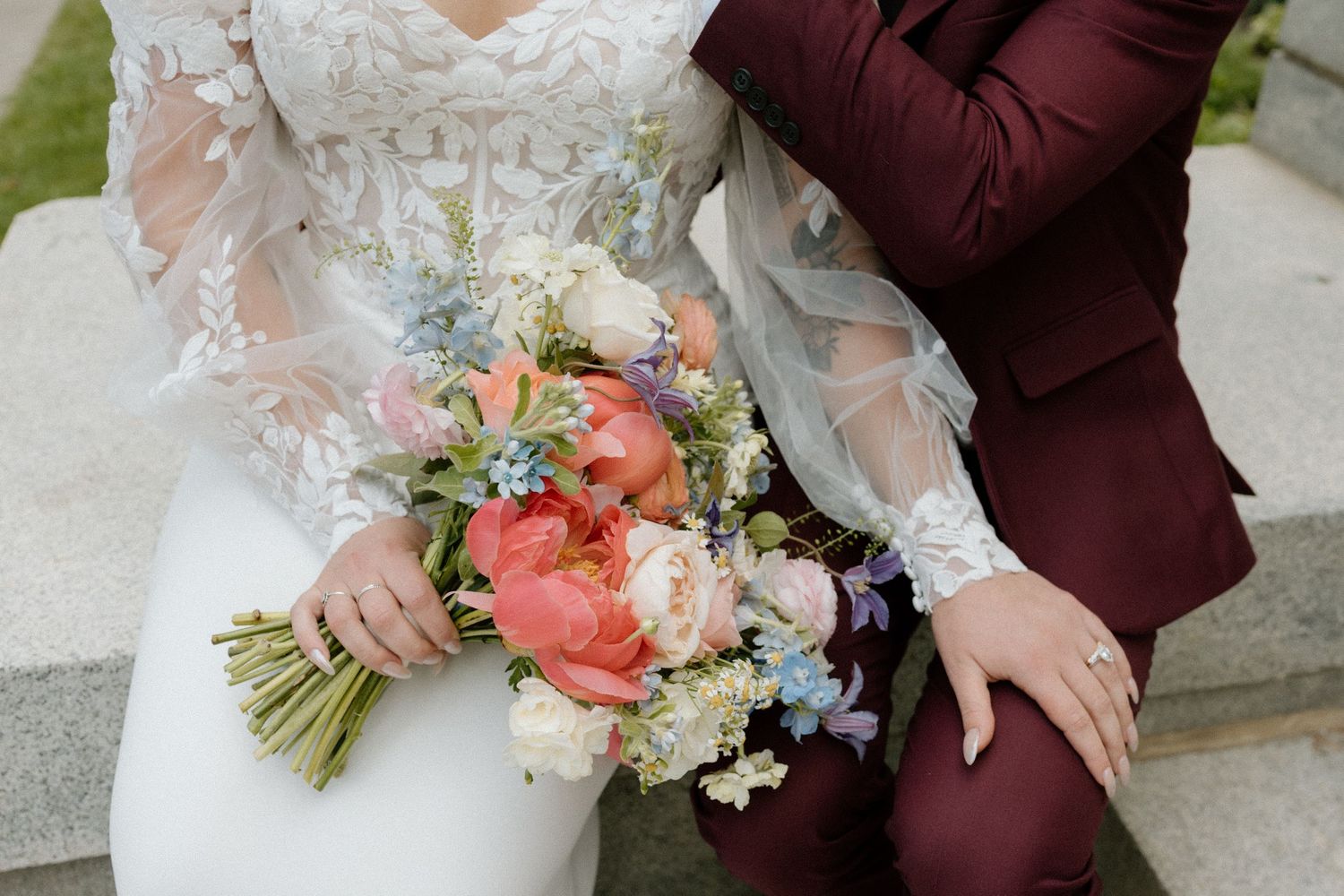 Close-up of wedding bouquet with coral and pastel flowers against white dress and burgundy suit.