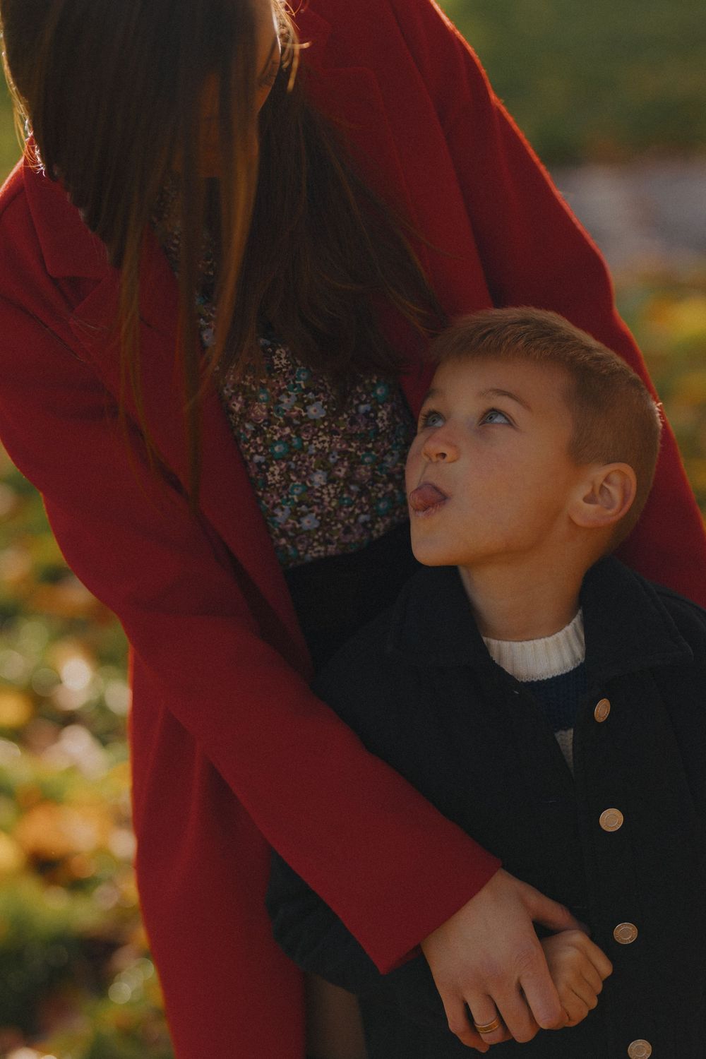 A child in a dark coat looks up at someone wearing a burgundy jacket against an autumn background.