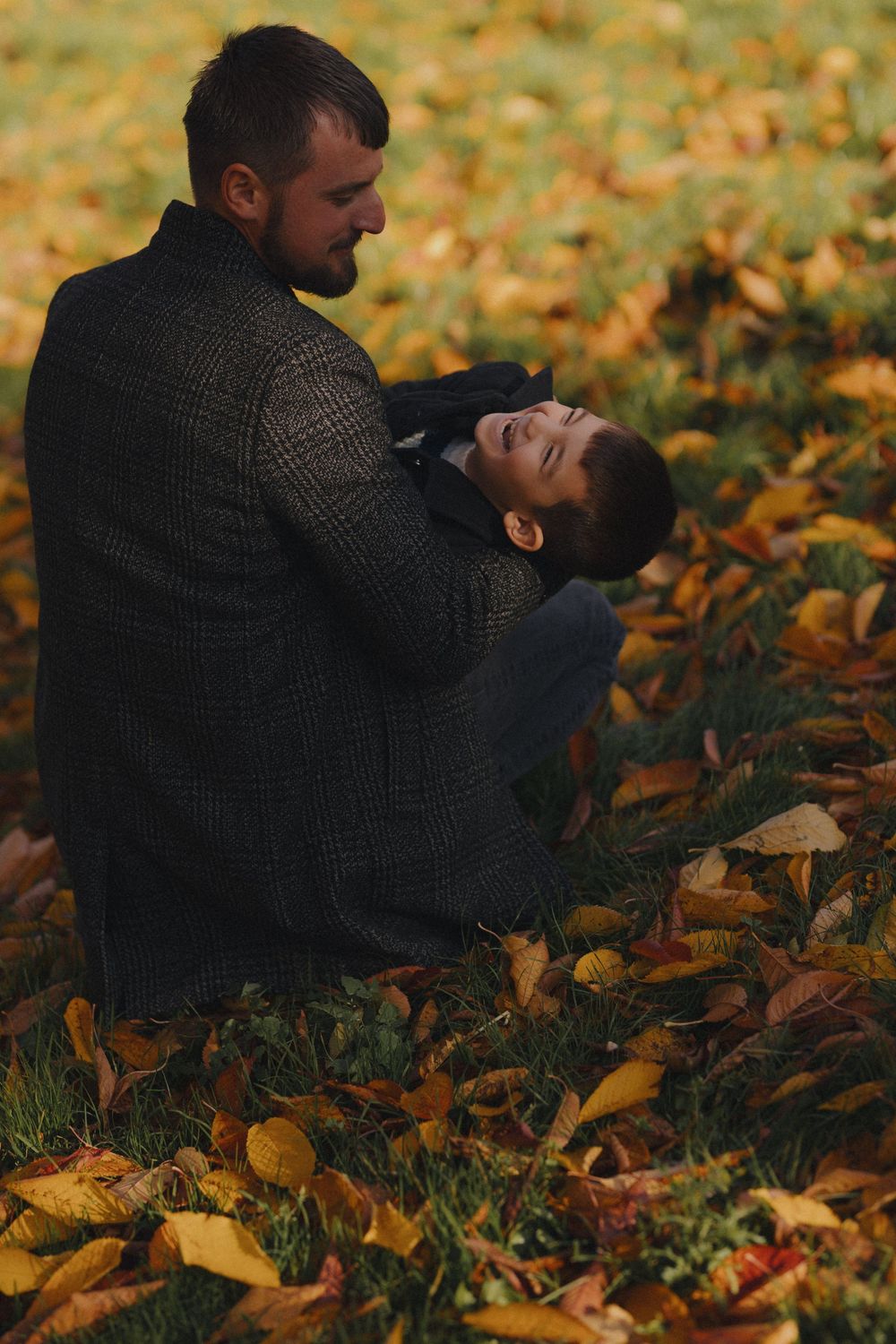A parent and child play together while lying in fallen autumn leaves on the ground.