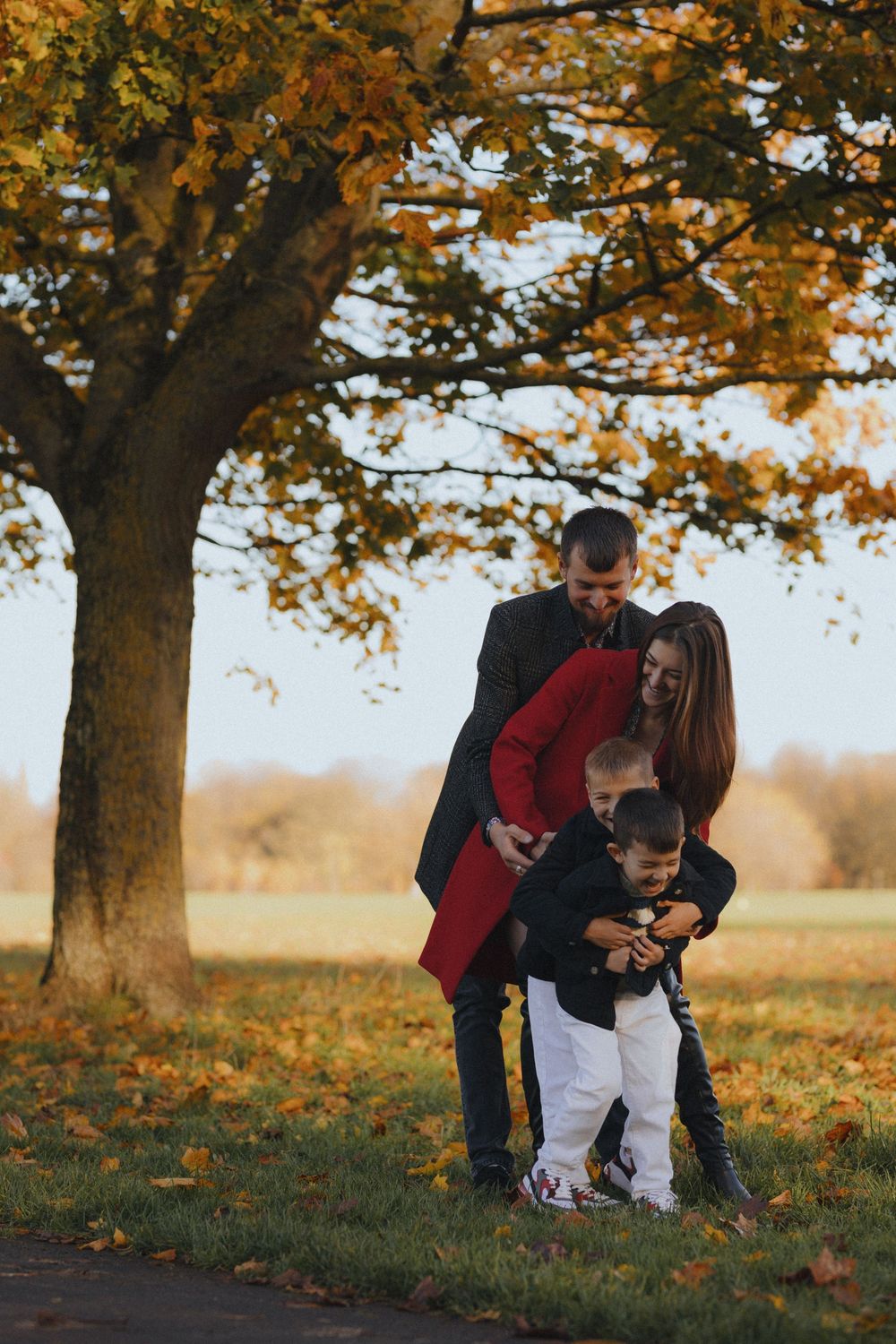 A family shares tender moments under autumn trees with golden leaves falling in a peaceful outdoor setting.