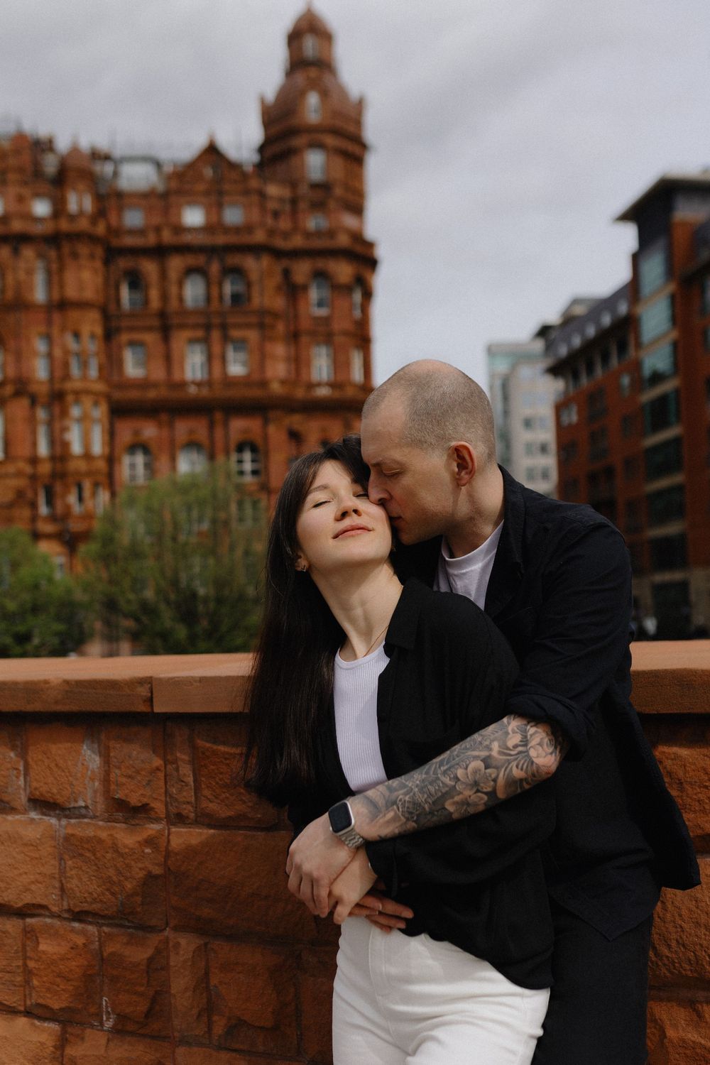 A couple embraces against a backdrop of historic red brick architecture in an urban setting.