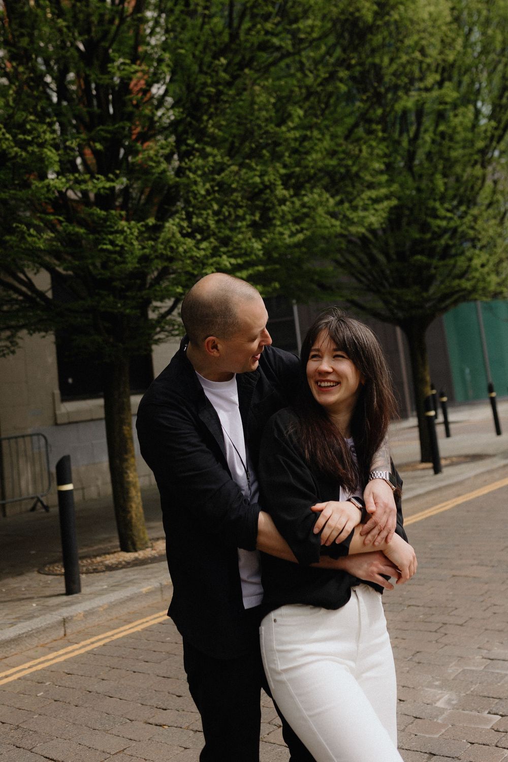A couple in contrasting black and white outfits share intimate moments while walking along a tree-lined street.