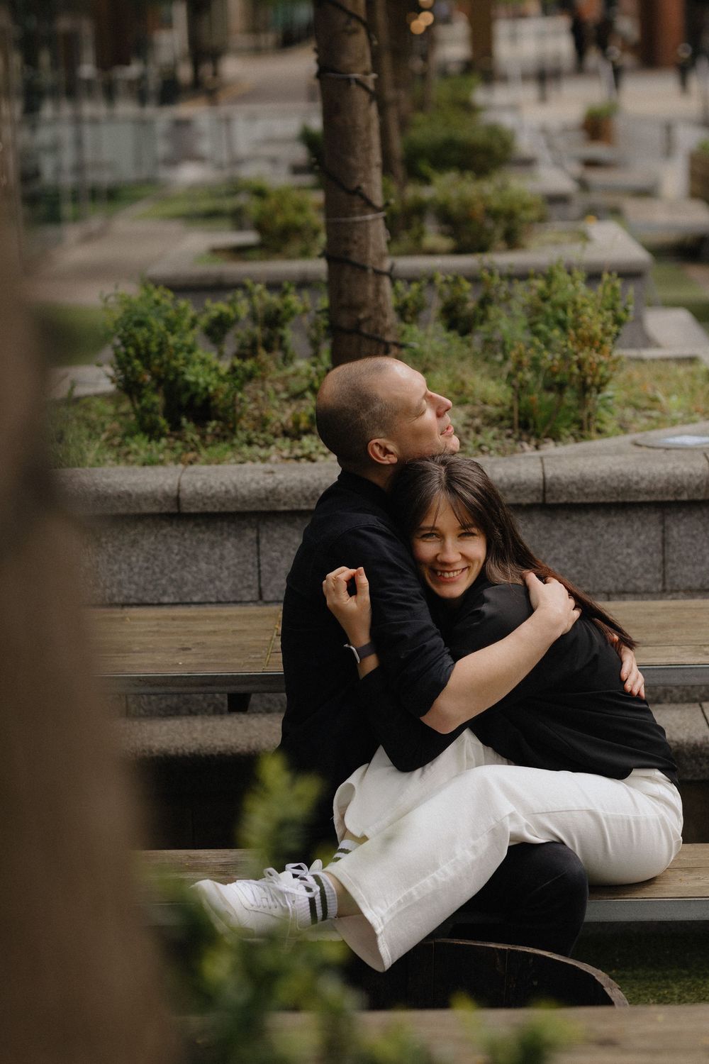 A couple shares a playful moment together while sitting on a stone bench in an urban park setting.