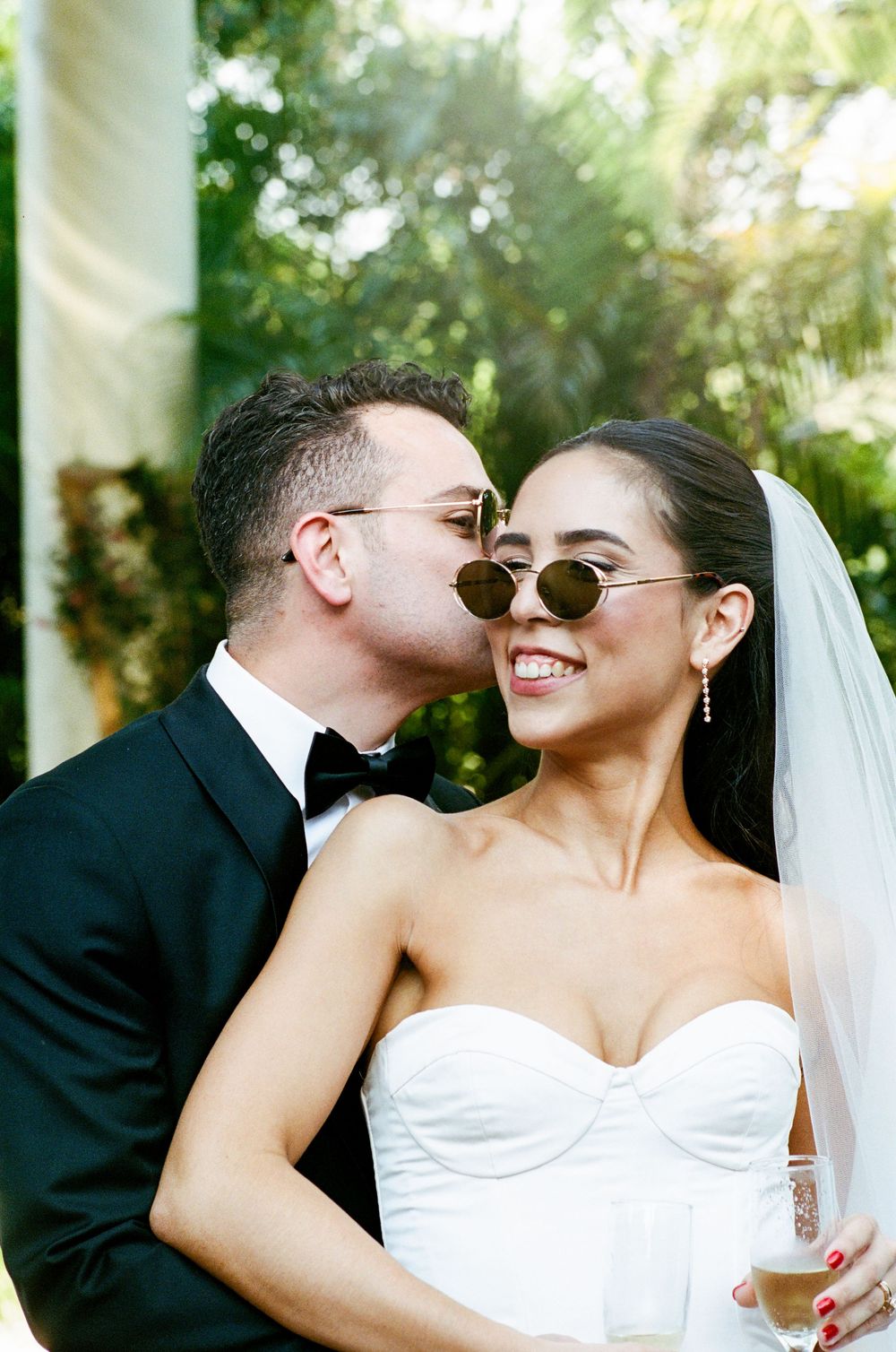 Groom in black tuxedo kissing bride wearing white strapless gown, veil, and sunglasses outdoors.