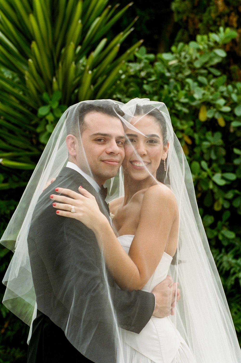 A happy couple shares intimate moments under a wedding veil against a lush green garden backdrop.