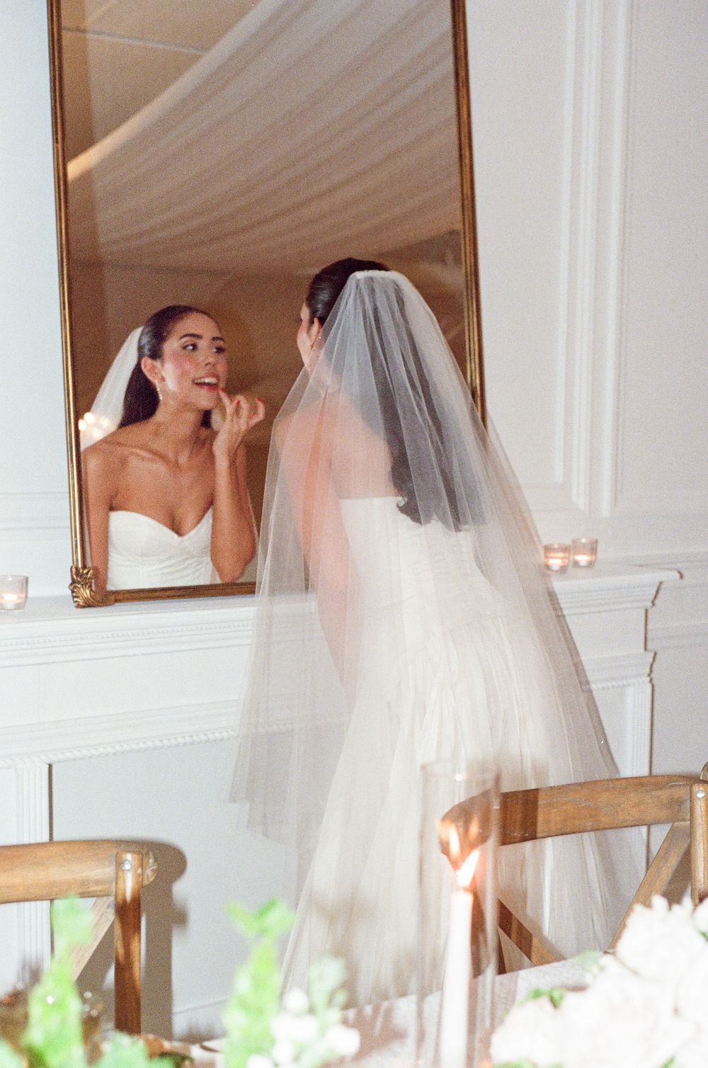A bride in a white wedding dress and veil is reflected multiple times in a mirrored wall above decorative greenery.