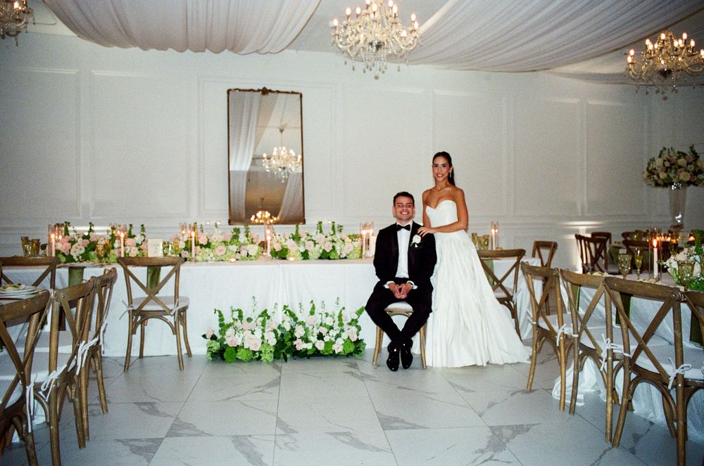 A panoramic view of an elegant white wedding reception venue with crystal chandeliers and crossback chairs.