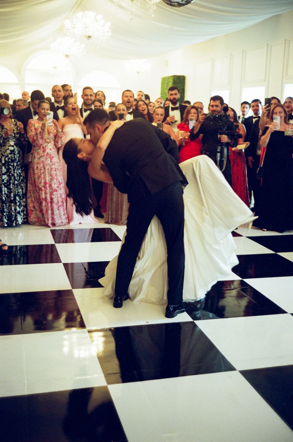 A couple performs their first dance at a wedding reception on a black and white checkered dance floor surrounded by guests.