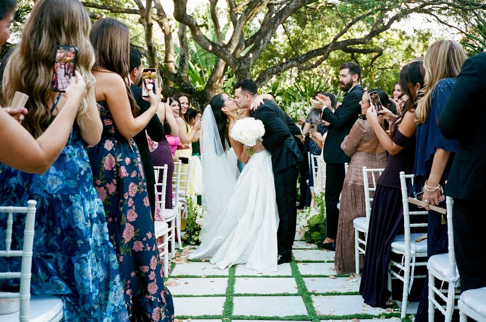 Wedding guests stand and celebrate during an outdoor ceremony under trees with white folding chairs.