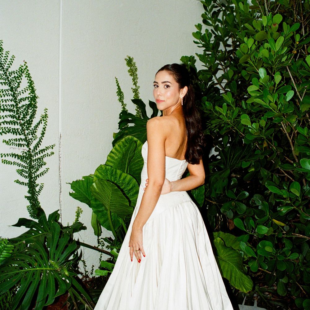 A woman in a white strapless gown poses against a backdrop of lush tropical greenery and ferns.