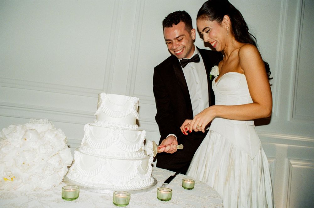 A couple in formal wedding attire cuts into an elegant white tiered wedding cake during their reception.