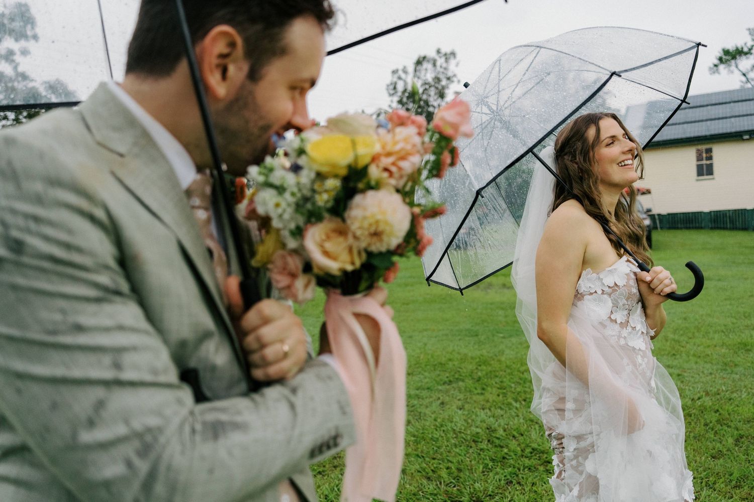 A romantic wedding couple shares tender moments under clear umbrellas in a lush green field on a rainy day.