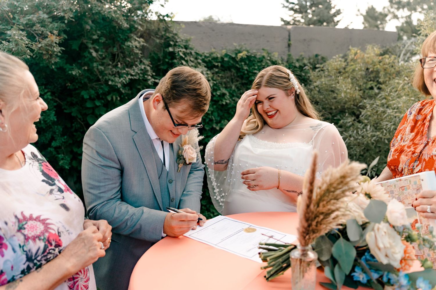 A couple in wedding attire signs documents at a round table with a peach-colored tablecloth during an outdoor ceremony.