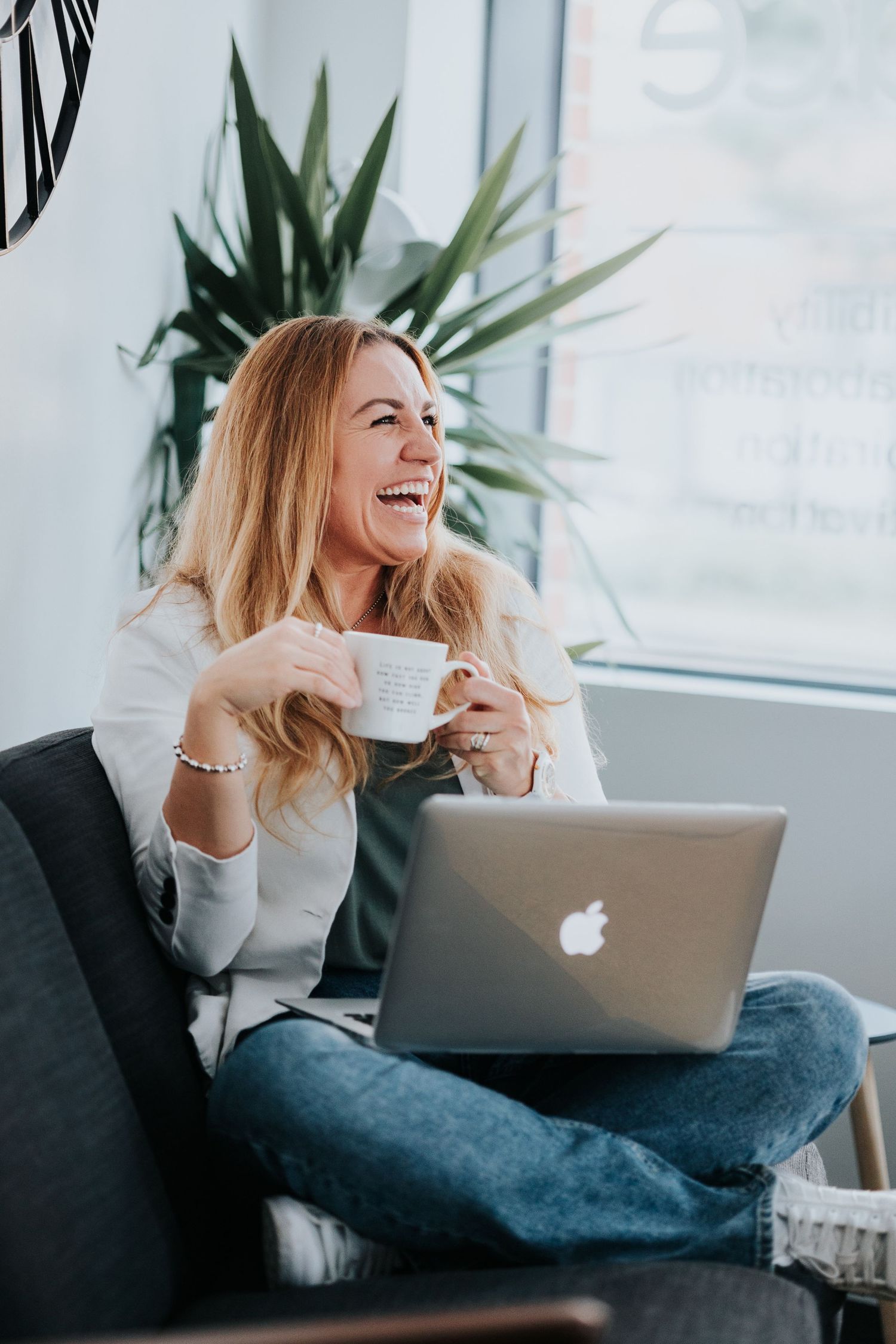 A person in a white sweater sits comfortably on a couch, working and laughing with a laptop in a bright room.