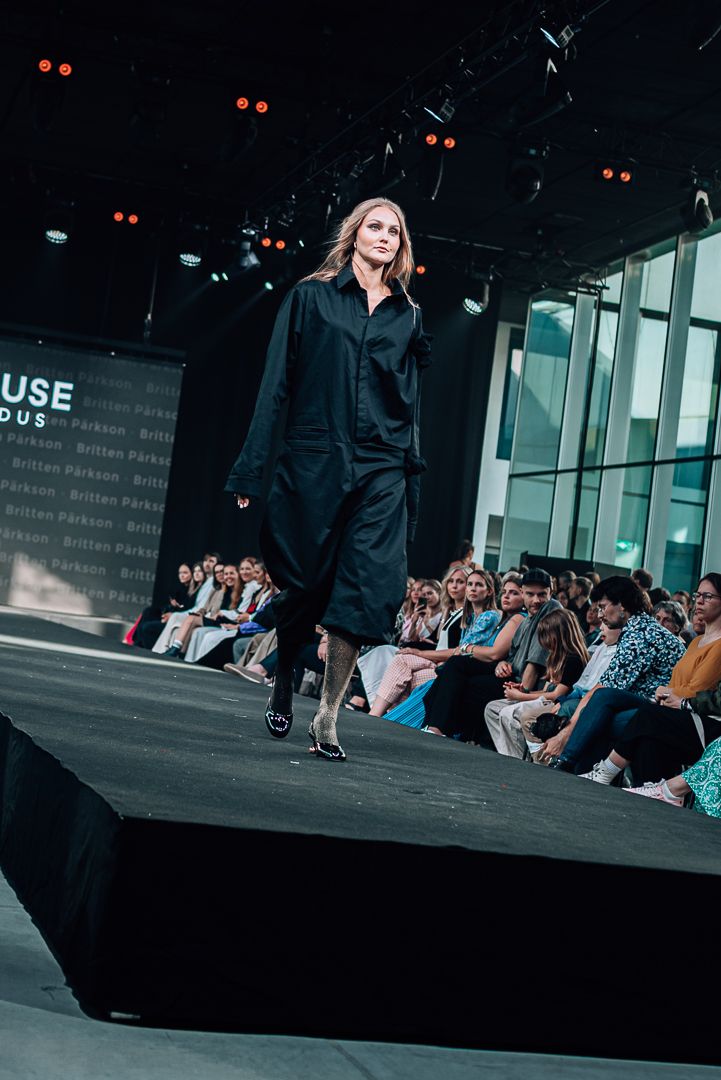 A model walks down a dark runway wearing an oversized black outfit during a fashion show with audience members seated.