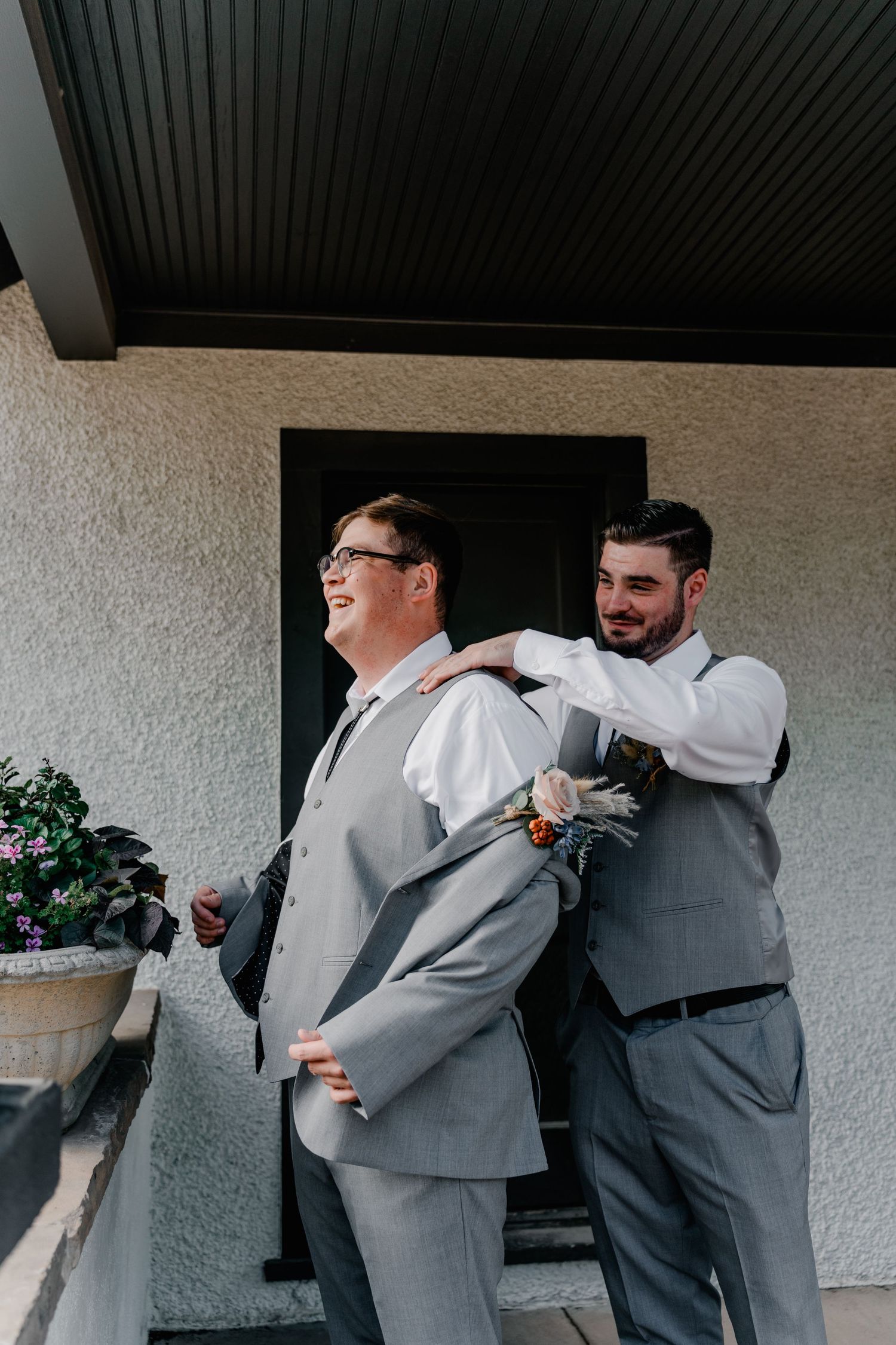 Two men in gray suits share a candid laughing moment while getting ready for a formal event.