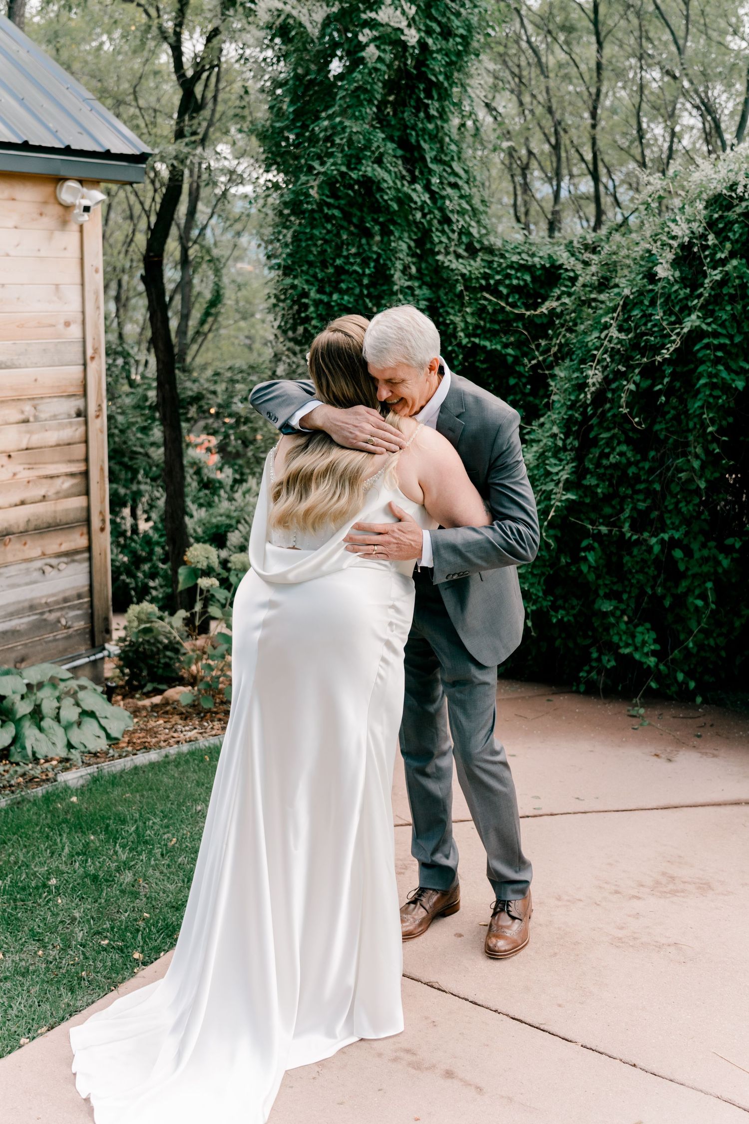 A bride in a white dress shares a joyful moment with loved ones outdoors near a rustic wooden wall.