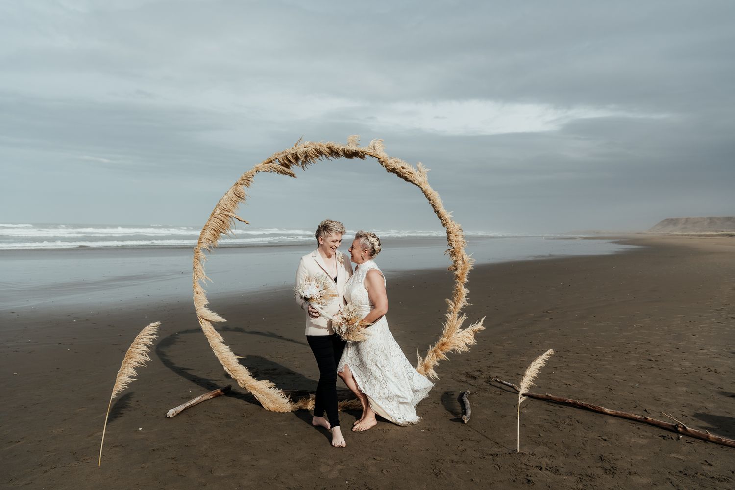 Under an overcast sky two brides stand on a deserted Riversdale beach with a circle arch of toi toi blowing in the wind by August & Vine