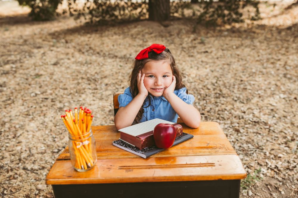 Student sitting at wooden desk with pencils and books in outdoor learning environment with dappled sunlight.