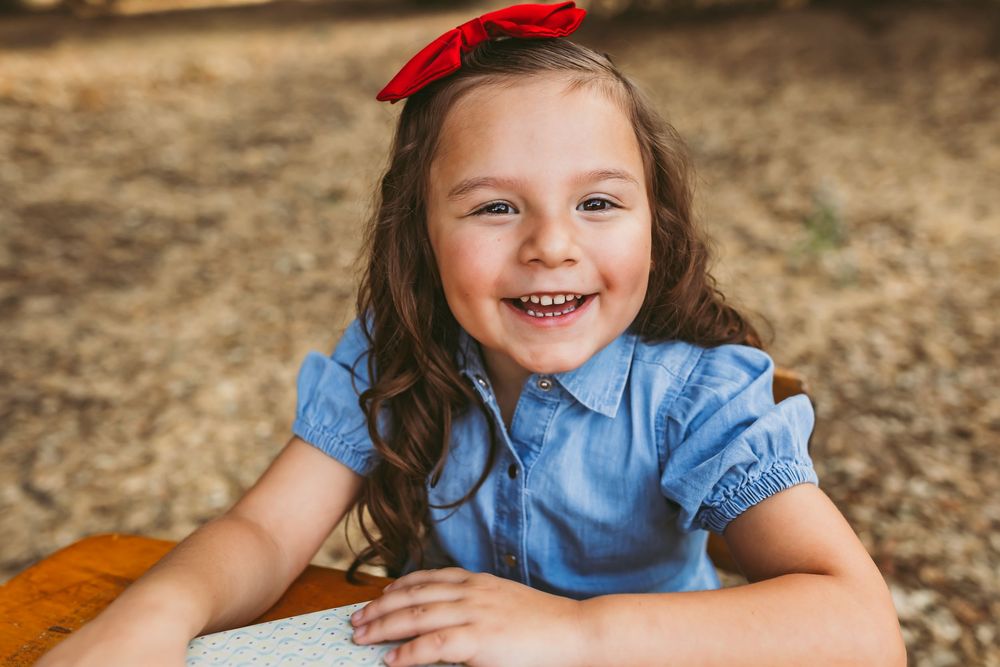 Young student wearing a blue dress and red bow shows excitement during back to school photo session outdoors.