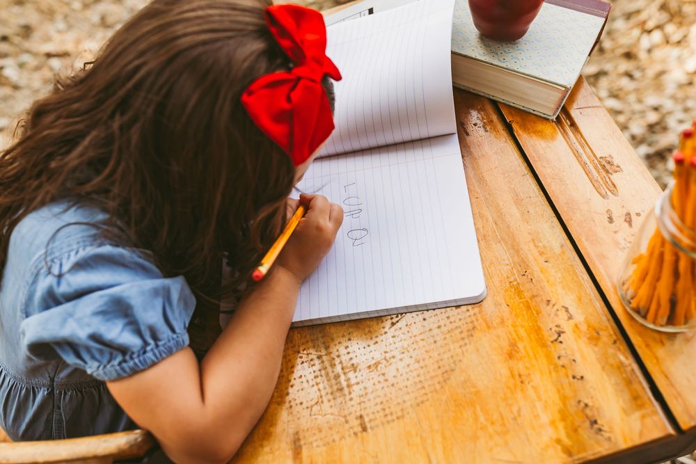Student with red hair bow works on writing in notebook at wooden desk outdoors.
