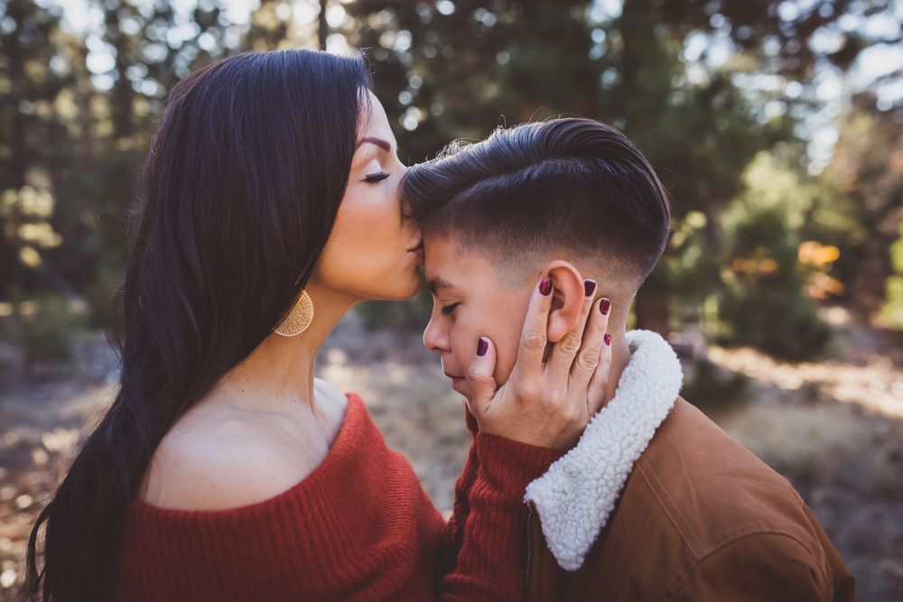 A couple embraces and kisses in a sunlit forest setting during a romantic outdoor photo session.