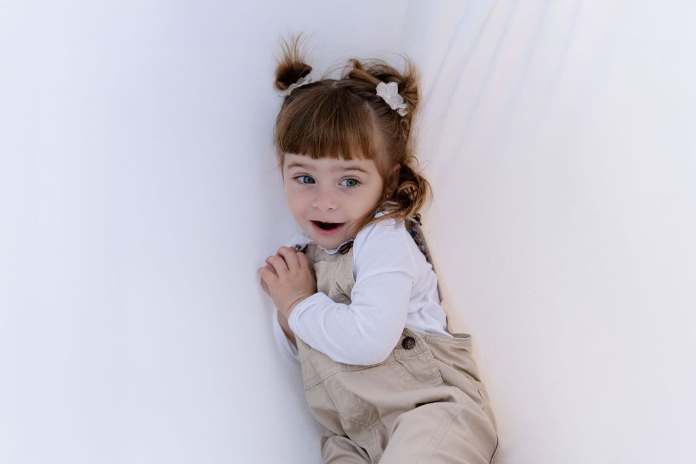 Child lying on light gray carpet wearing beige sweater and white ruffled collar looks up with joyful expression.
