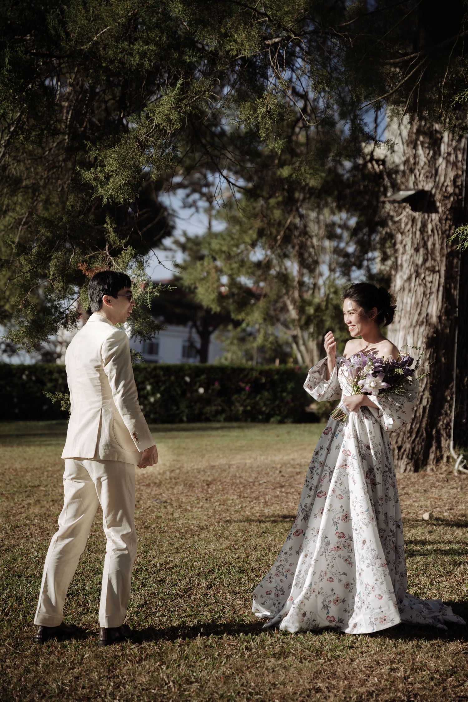 Bride in floral gown holding bouquet smiling at groom in cream suit beneath large pine tree.