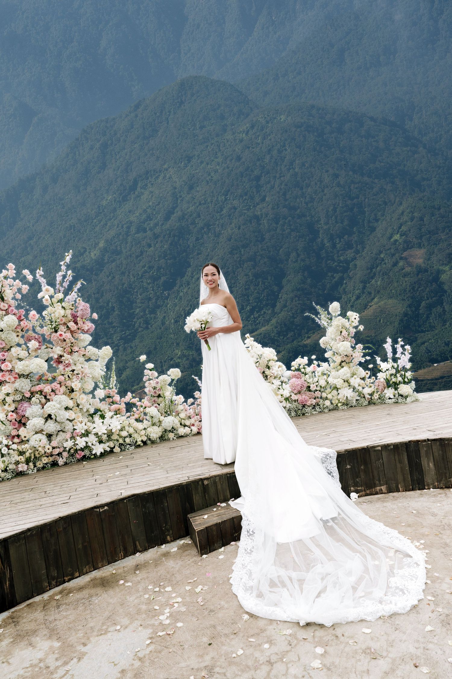 Bride in flowing white gown with long veil stands before pink and white floral arch with mountain backdrop.