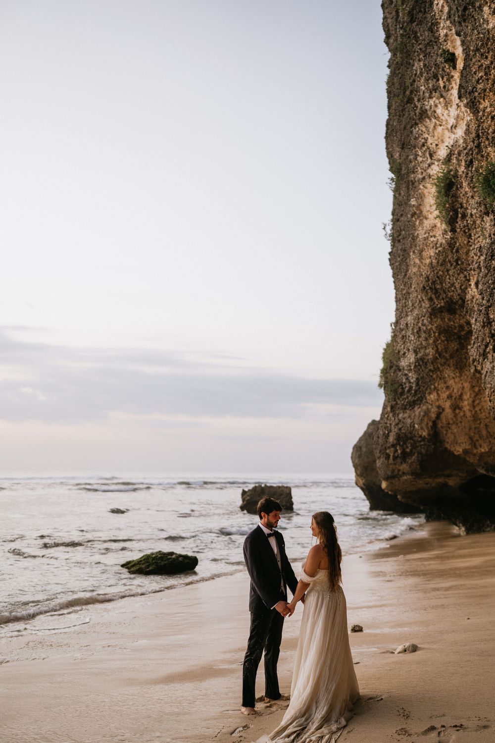 Elegant beach wedding portraits captured beneath towering coastal cliffs at sunset.