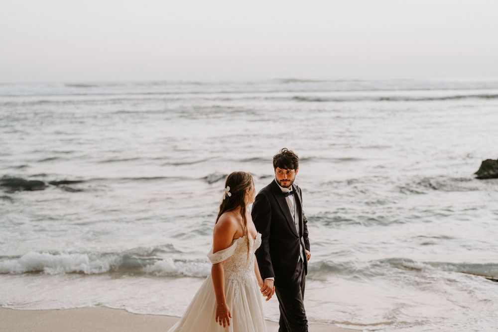 Couple in formal attire walking along rocky coastline during overcast day.