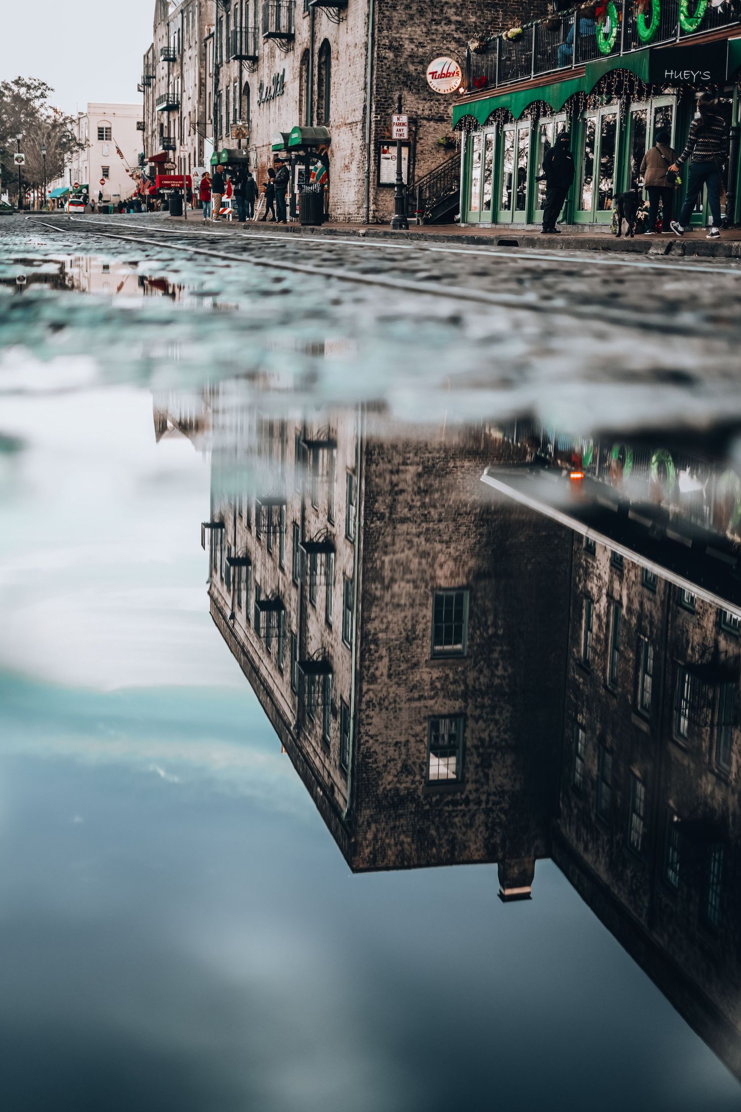 Urban street scene reflected in puddle with stone buildings and storefront signs on a cloudy day.