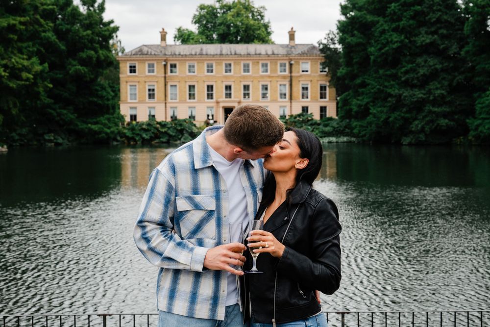 A romantic moment shared between two people standing by a lake with a grand English manor house in the background.