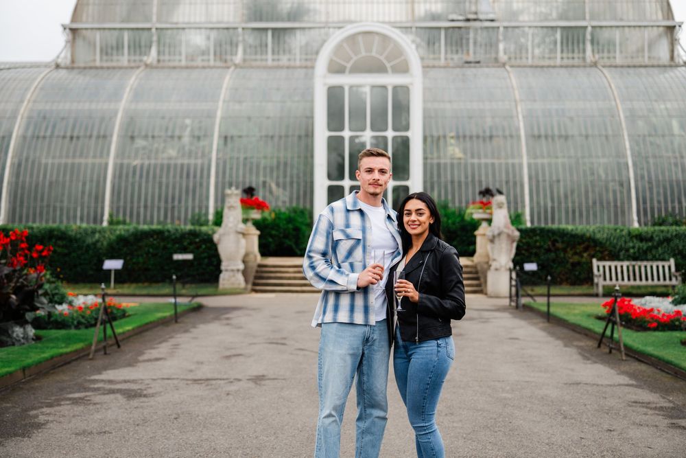 A couple stands in front of a grand glass conservatory with ornate architecture and red flowers along the walkway.
