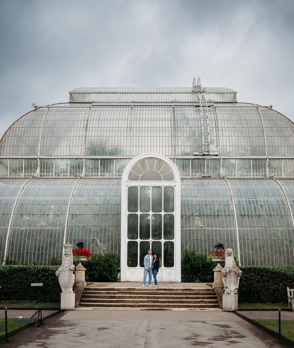 A grand Victorian glasshouse greenhouse with a curved glass dome and arched entrance features ornate stone statues at its steps.