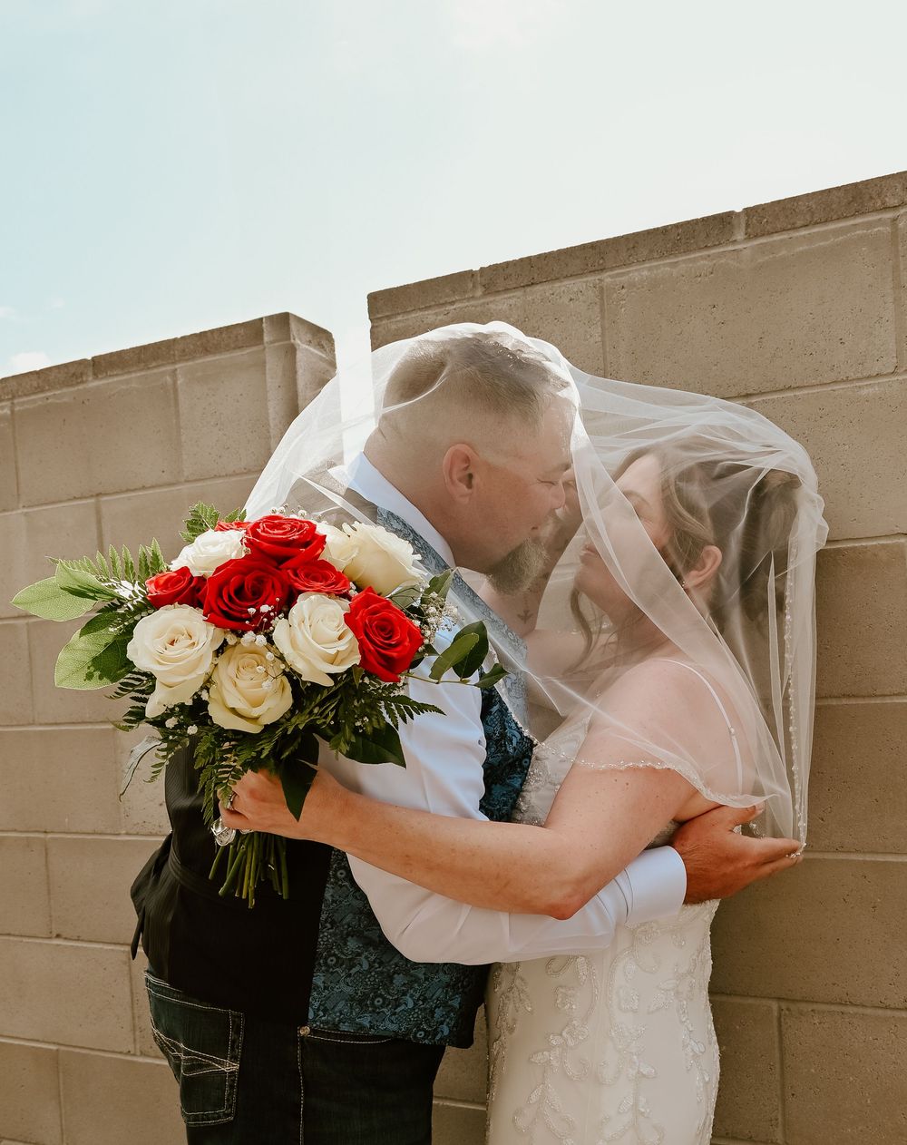 Wedding couple embraces in an outdoor setting with elegant floral bouquets of red and white roses.