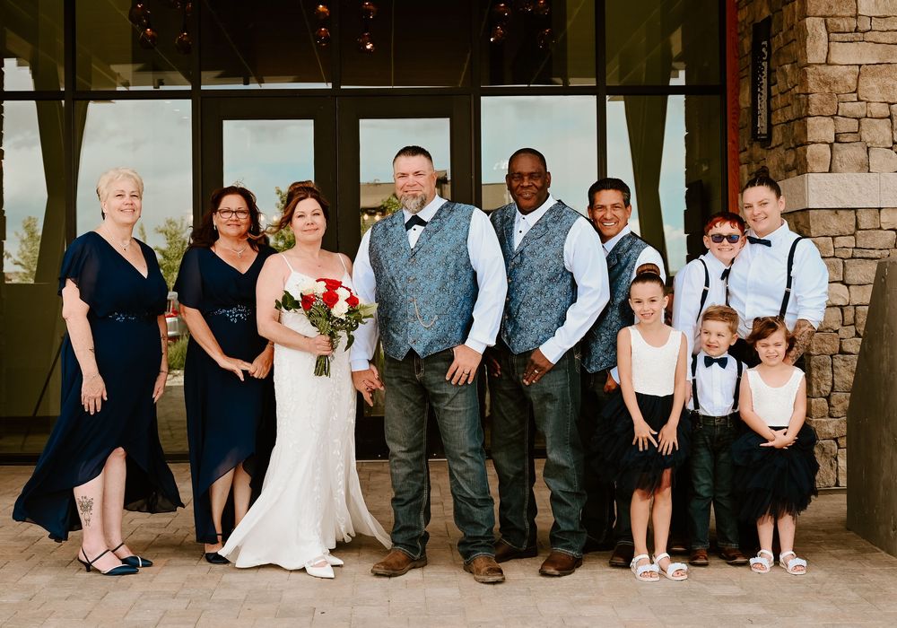 A wedding party group poses together outside a stone building with glass windows on a sunny day.