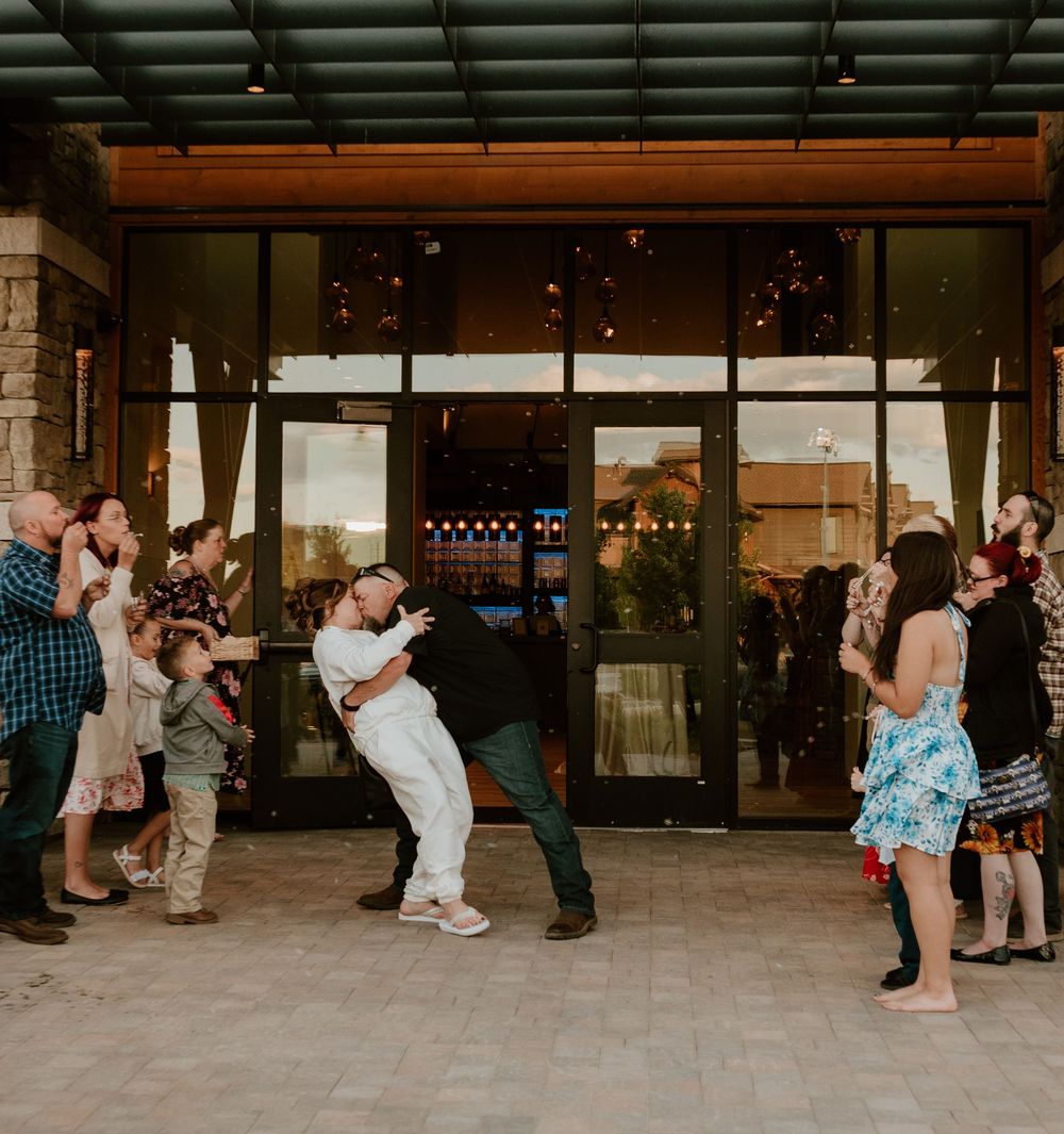 Wedding guests gather outside a modern venue entrance with glass doors and string lights visible inside.