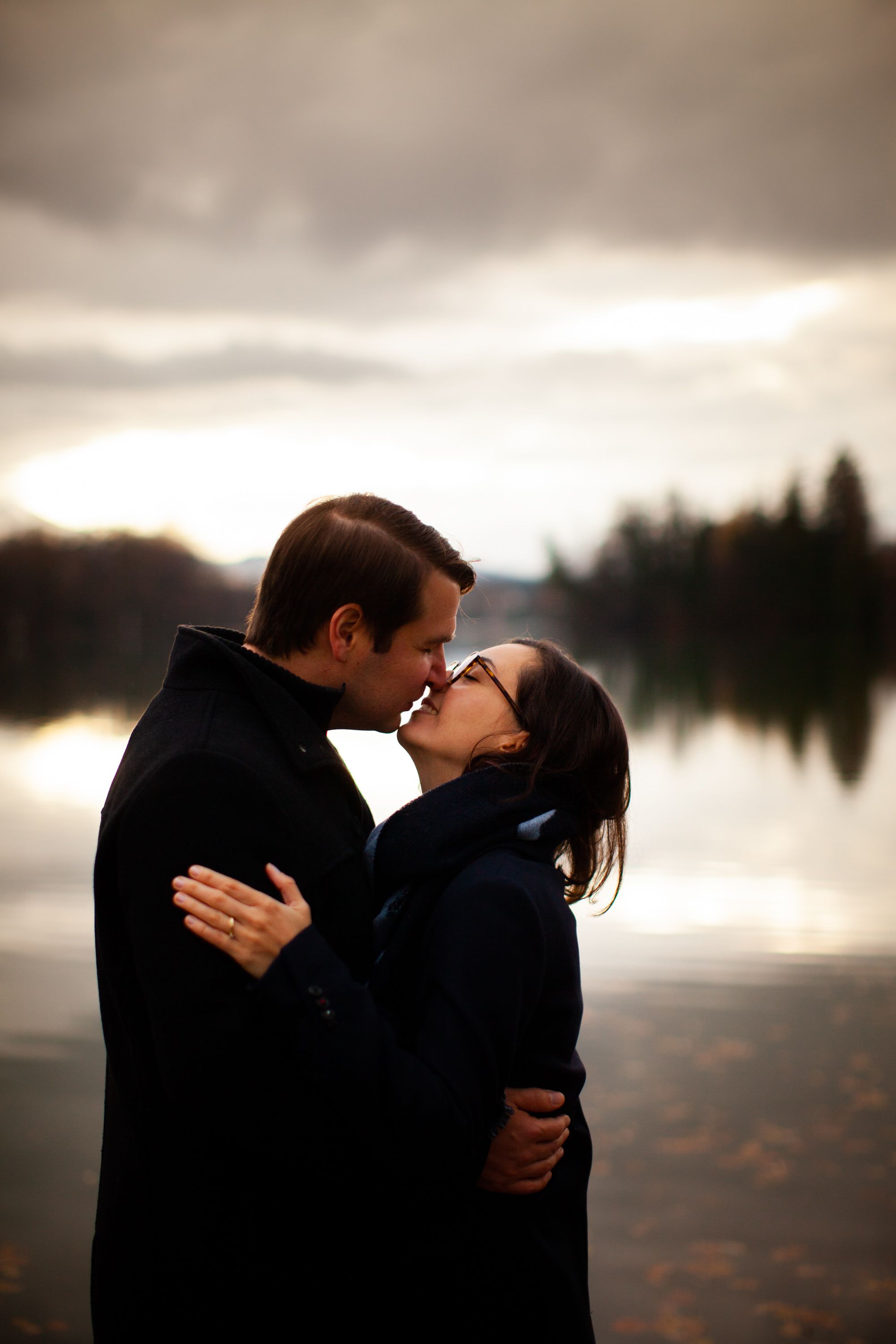 A romantic silhouette of a couple sharing a kiss at sunset by a tranquil lake with foggy mountains in the background.