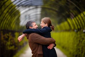 A couple embraces in a romantic moment under an arched tunnel of green foliage in a garden setting.